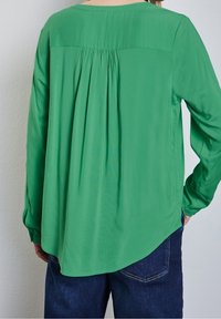 Camicia verde a maniche lunghe con schiena plissè, scollo rotondo, tessuto leggero e orlo curvo. Abbinata a jeans blu scuro.