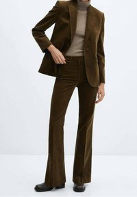 Ruskea keinonahkainen bleiseri istuvalla leikkauksella, yhdistetty samaan sarjaan kuuluviin lievästi levenevään housuihin. Poolopusero on vaalean beige, mikä luo kontrastia.