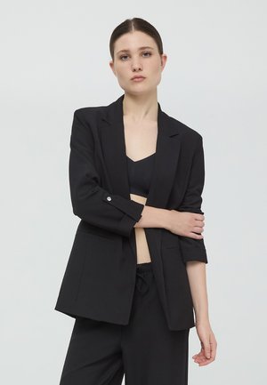 Blazer nero con vestibilità rilassata, design a un bottone, due tasche frontali e maniche arrotolate. Indossato sopra una maglia nera.