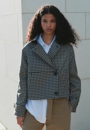 Kariertes kurz geschnittenes Jackett aus grün-cremigem Stoff, mit weitem Kragen, zwei Vorderentaschen und Knopfverschluss, über einem weißen Hemd getragen.