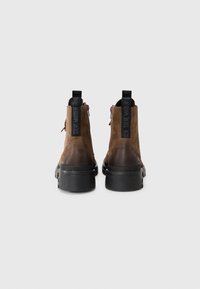 Brown suede ankle boots with a textured finish, black rubber sole, and contrasting black pull tab featuring logo detail. Side zip closure.