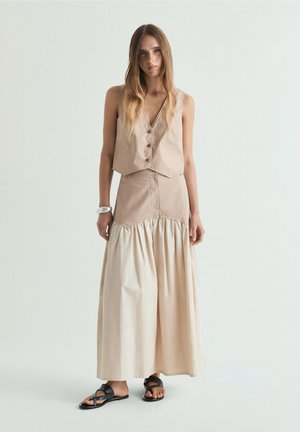 Femme aux cheveux longs portant un gilet beige sans manches et une longue jupe bicolore assortie, debout avec un bras légèrement plié et des sandales noires.