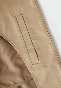 Lys brun jakke stof med en glat tekstur, der har en lynlåsåbning og en sidelomme syet på forsiden.