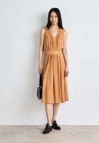Une robe midi sans manches, décolletée en V, en tissu orange clair avec des plis, une taille cintrée accentuée par des coutures décoratives, associée à des chaussures noires.
