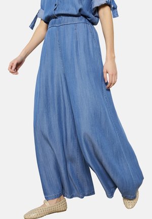 Femme portant un pantalon en denim bleu clair, large et ample, associé à des chaussures plates beiges cloutées, debout devant un fond uni.