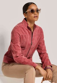 Rote Wildlederjacke mit Reißverschluss, gequilteten Paneelen und langen Ärmeln. Braune, schmale Hose. Klare, übergroße Sonnenbrille mit leichtem Farbton.