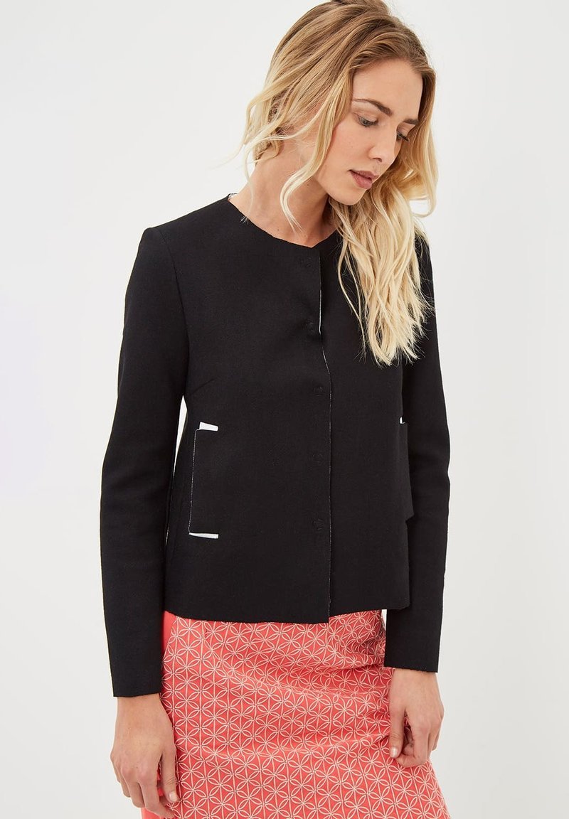 Blazer negro con cuello redondo, con ribete blanco en los bolsillos y al frente. Tejido liso, diseño ajustado, combinado con una falda rosa estampada.