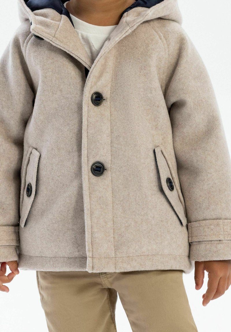 Enfant portant un manteau en laine beige boutonné avec des poches, par-dessus un pull clair et un pantalon beige, debout sur fond blanc.