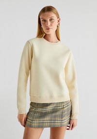 Crème sweatshirt met ribbels rond de hals en mouwen, gecombineerd met een geruite rok in gele, blauwe en zwarte kleuren; over het algemeen een gladde textuur.