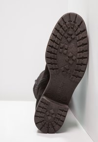 Semelle d'une botte noire avec des profondeurs de semelle montrant des motifs d'adhérence et le nom de la marque "Gabor", inclinée contre une surface et un mur blancs.