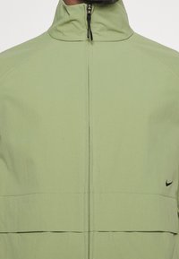 Chaqueta verde claro con cremallera y alto cuello, diseño elegante, con el logo negro de Nike en la parte inferior izquierda. Textura suave y detalles sin costuras.