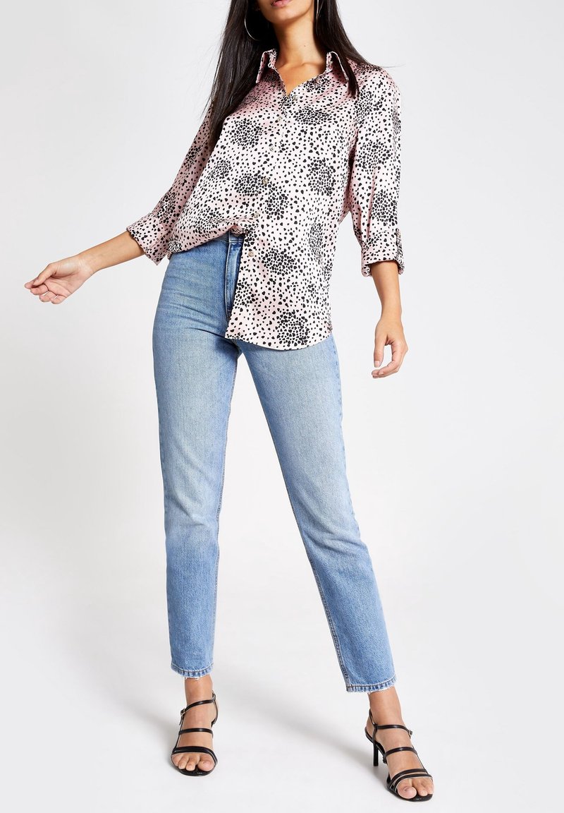 Femme portant une chemise à manches longues rose et noire à pois, un jean bleu clair et des sandales à talons hauts noires à brides, debout devant un fond blanc.