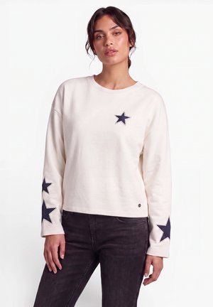 Donna che indossa una camicia bianca a maniche lunghe con toppe a forma di stella blu navy sul petto e sulle maniche, abbinata a jeans scuri, in piedi su uno sfondo bianco.