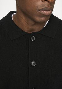 Sort strikket cardigan med krave, der har fire sorte knapper og en struktureret stof. Hvid skjorte synlig nedenunder.