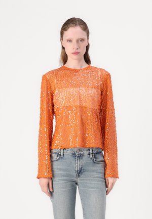 Oransje langermet topp laget av gjennomsiktig stoff dekket med paljetter, med rund hals og croppet passform. Kombinert med lyseblå denimjeans.