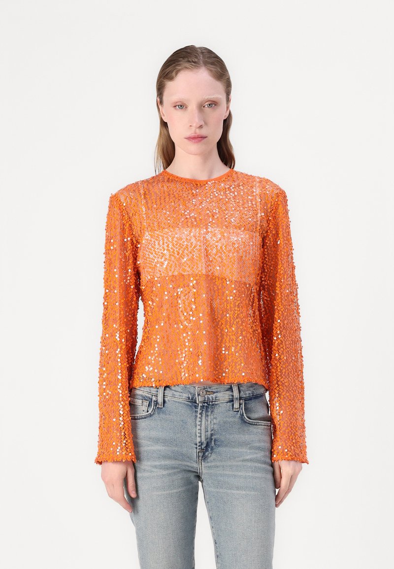 Oranje lange mouwen top van doorzichtig materiaal bedekt met pailletten, met een ronde hals en een cropped fit. Gecombineerd met lichtblauwe denim jeans.