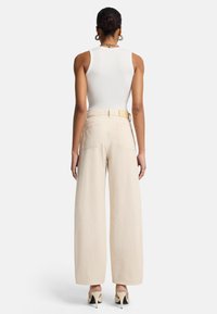Beige weite Hose mit hoher Taille und Gesäßtaschen, kombiniert mit einem weißen figurbetonten Tanktop und hohen Schuhen, von hinten präsentiert.