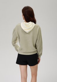 Fleece mikina s kapucí ve světle zelené barvě s krémovým vnitřkem kapuce, s volným střihem a dlouhými rukávy, v kombinaci s černými denimovými šortkami.