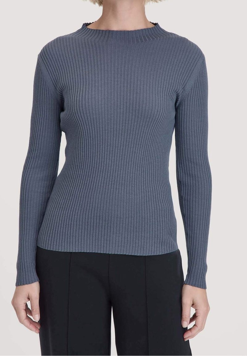 Femme portant un pull gris ajusté à manches longues, côtelé, associé à un pantalon noir ample, vue du cou jusqu’aux hanches sur un fond uni.