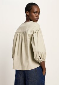 Schwarze Frau mit geflochtenem Haar, die eine lockere beige Bluse und blaue Denim-Hosen trägt, schaut über ihre Schulter vor einem schlichten Hintergrund.