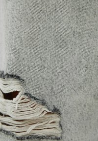 Vaaleanharmaa farkkukangas, jossa on vaaka suuntaan kulkeva repeämä, joka paljastaa alapuolella olevia kerroksellisia valkoisia langoja vasemmassa alakulmassa.