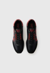 Schwarze Sportschuhe mit roten Akzenten und Logo, ausgestattet mit einem glatten synthetischen Obermaterial, Schnürverschluss und einer kontrastierenden weißen Gummisohle.