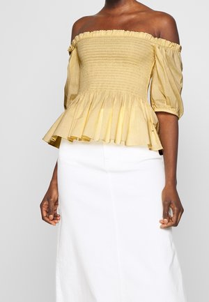 Femme portant un haut jaune à épaules dénudées avec manches bouffantes et un pantalon blanc taille haute, sur un fond uni.