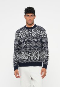 Granatowy sweter dzianinowy z białym wzorem geometrycznym, okrągłym dekoltem oraz ściągaczami przy mankietach i u dołu, noszony na jasnych spodniach.
