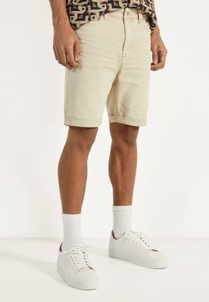 Mann trägt beige Shorts mit umgekrempeltem Saum, weiße Socken, weiße Turnschuhe und ein gemustertes Hemd, steht vor einem schlichten Hintergrund.