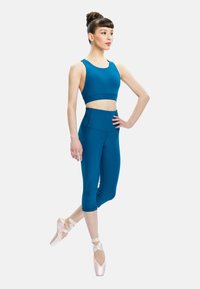 Female ballet dancer in teal crop top and leggings poses on pointe shoes against plain white background.