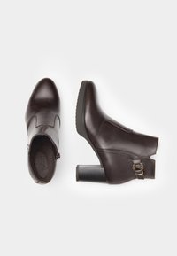Stivaletti in pelle marrone fino alla caviglia con punta affusolata, chiusura con cerniera e tacco grosso. Presentano un design con ritaglio e un accento in fibbia dorata.