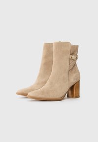 Stivali ankle in suede beige con punta affusolata, tacco in legno impilato e una fascia con dettaglio a fibbia dorata sul lato.