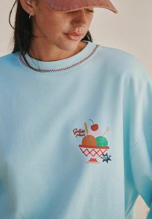 Woman wearing a light blue sweatshirt with a colorful ice cream bowl and "Gelato please" text embroidered on the chest.