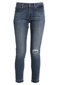 Jeans ajustados de mezclilla azul oscura, con un desgastado en la rodilla derecha y un diseño estándar de cinco bolsillos con cierre de cremallera.
