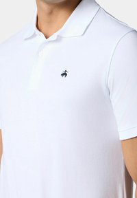 Bílá polo košile vyrobená z hladké tkaniny, s klasickým límečkem a námořnickomodrou vyšitou logem na levém prsu.