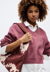 Γυναίκα με σγουρά μαλλιά φοράει βυσσινί πουλόβερ με V-λαιμόκοψη πάνω από λευκό πουκάμισο, κρατώντας μια μεγάλη τσάντα ώμου με καφέ και λευκό σχέδιο.