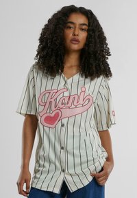 Baseballstil skjorte i kremfarge med svarte striper. Har en stor rosa grafisk tekst "Kani" og hjerte-design. Korte ermer, knapp foran.