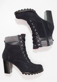 Tmavě modré semišové kotníkové boty s robustním podpatkem, vybavené šněrováním, bočním zipem a texturovaným lemem pro zvýraznění detailů a lepší podporu.