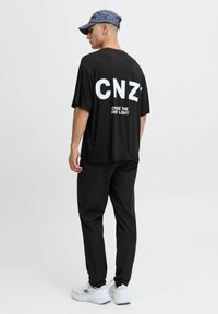 Camiseta negra de gran tamaño con letras blancas "CNZ" y el texto "PERSEGUIDE LA PRIMERA LUZ" en la parte posterior, combinada con pantalones negros y zapatillas blancas.