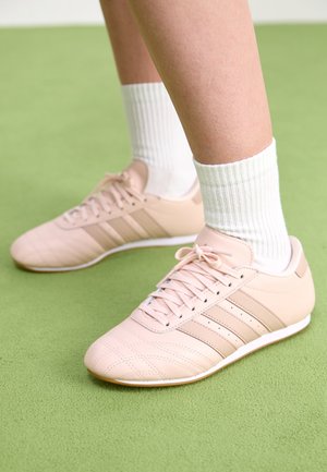 Pieds portant des baskets Adidas rose clair avec semelles blanches et des chaussettes blanches côtelées, debout sur un tapis vert.