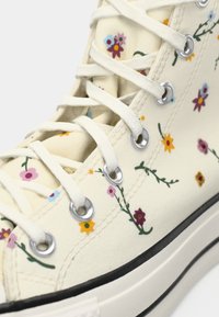 Sapatilhas altas em lona creme com estampa floral colorida. Apresentam ilhós prateados, atacadores brancos e sola de borracha preta.