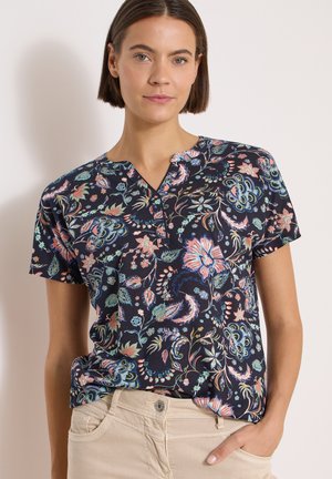Femme aux cheveux courts et bruns portant un chemisier bleu marine à fleurs à manches courtes et un pantalon beige, debout avec une main dans la poche.