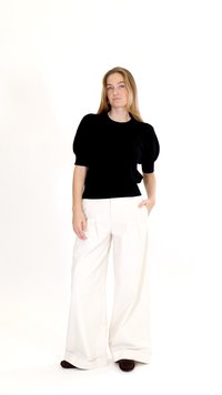 Frau steht mit einer Hand in der Tasche, trägt ein schwarzes Oberteil mit Puffärmeln, weiße Hose mit weitem Bein und dunkle Schuhe vor einem weißen Hintergrund.