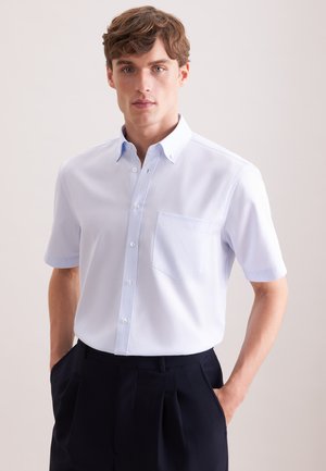 Młody mężczyzna ubrany w jasnoniebieską, krótko rękawową koszulę na guziki z kieszenią na piersi oraz czarne spodnie z plisami, ręce w kieszeniach.