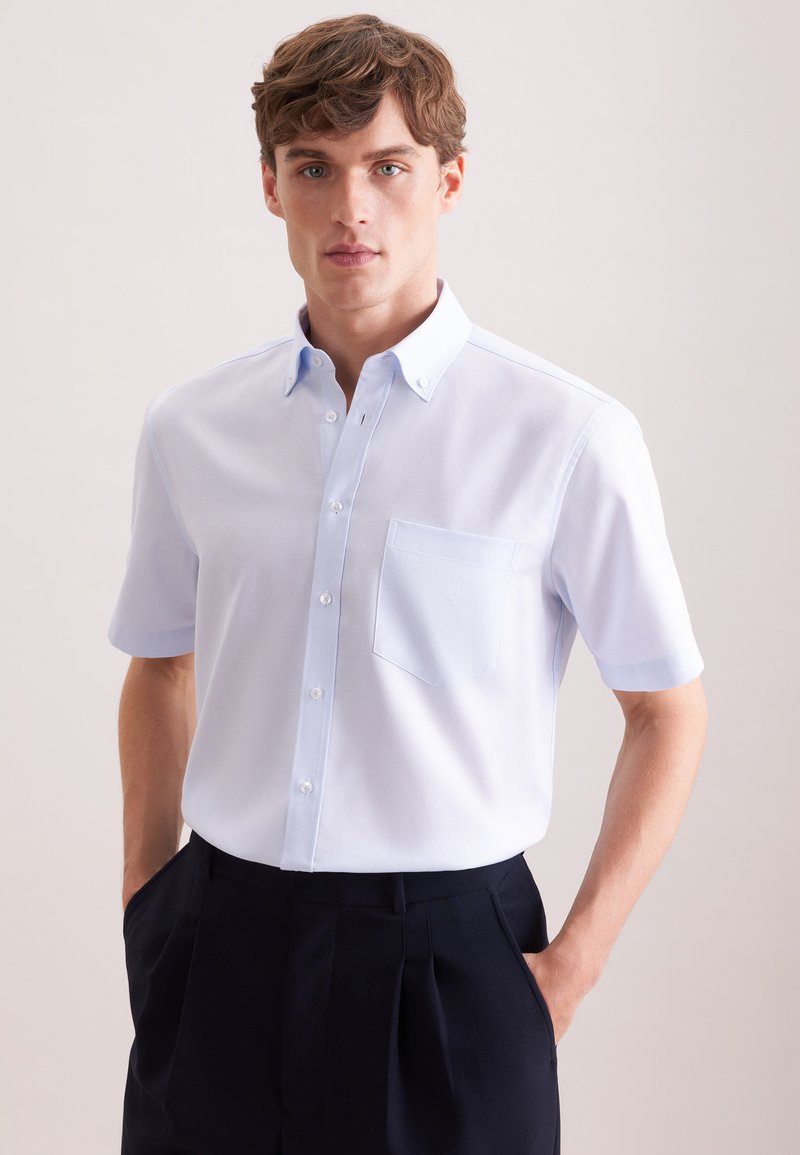 Jonge man die een lichtblauw overhemd met korte mouwen en een borstzak draagt, met zwarte geplooide broek en handen in de zakken.