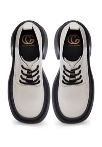 Pantofi din piele albă lustruită, cu șireturi negre și o talpă groasă neagră. Prezintă un logo auriu pe branțuri. Textură netedă, formă rotundă.
