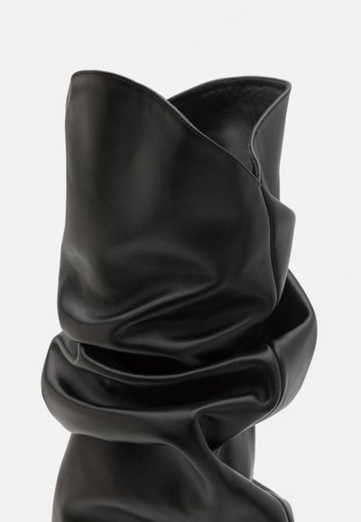 Botină din piele neagră până la gleznă, cu o textură moale și șifonată, și un guler înalt, asimetric. Prezintă un finisaj neted și un design minimalist.