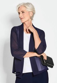 Marine sheer kimono jas met driekwart mouwen, losse pasvorm, gedragen over een witte top, en houdt een zwarte clutch met metallic accenten vast.