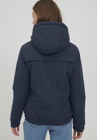 Giacca imbottita blu navy con cappuccio staccabile, polsini e orlo elastici; texture liscia con un leggero lucido; design minimalista.