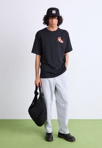 T-shirt noir avec graphique sur le devant, jeans bleu clair, sac matelassé noir et bottes noires épaisses. Le mannequin se tient sur un sol vert devant un fond clair.